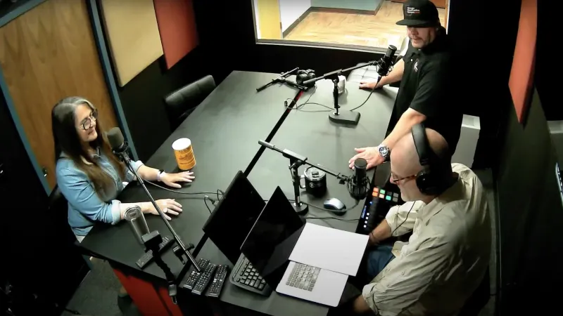 Three people recording a podcast in a soundproof studio with microphones, laptops, and headphones.
