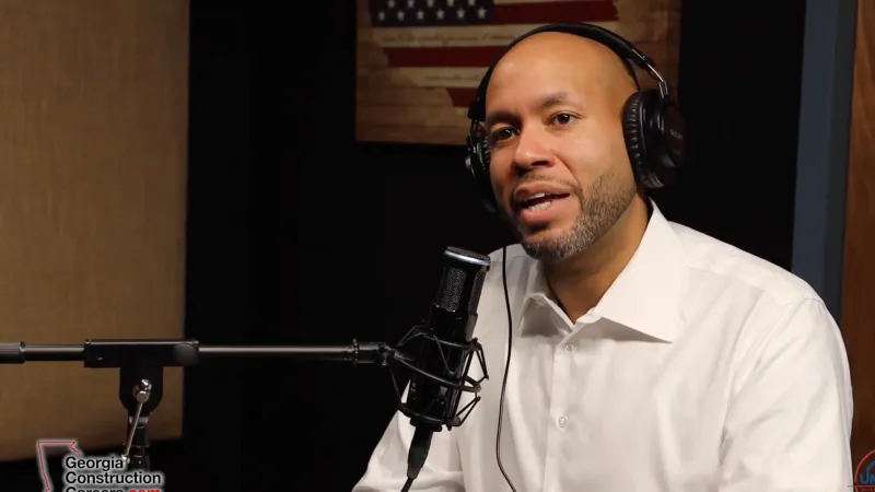 Man wearing headphones speaking into a microphone during a podcast recording with an American flag in the background.