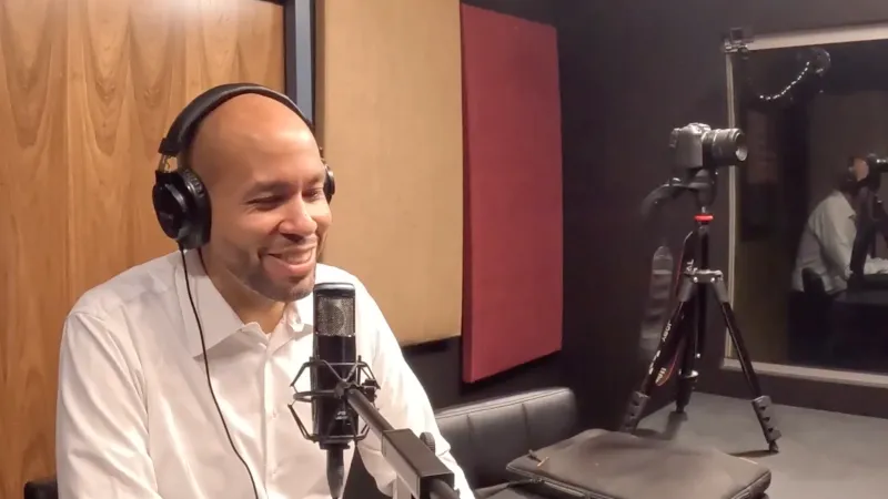 Man in white shirt with headphones speaking into a microphone in a podcast studio with acoustic panels.