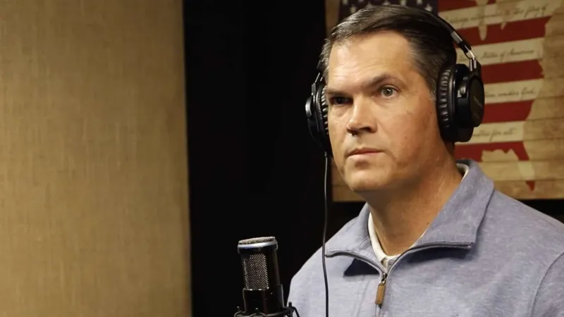 Man wearing headphones speaking into a microphone with a USA flag wall art in the background in a podcast studio