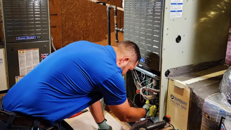 Technician in blue shirt repairing HVAC furnace system in a utility room with tools and equipment nearby.