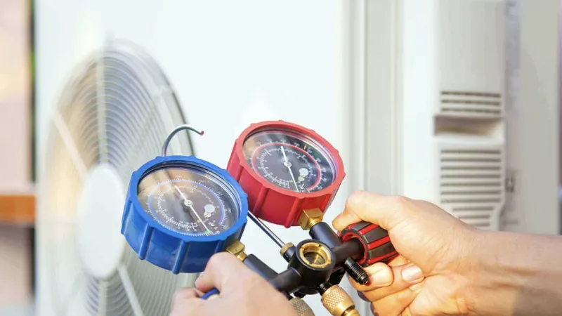 Technician holds HVAC manifold gauges with red and blue dials near air conditioning unit for pressure check