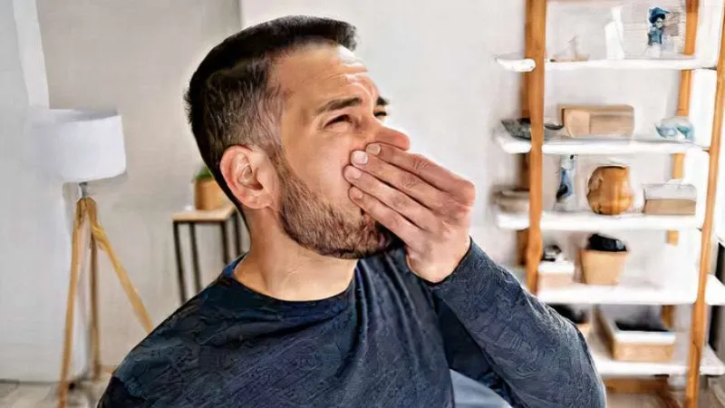 Man yawning and covering his mouth in a cozy room with shelves and a lamp in the background