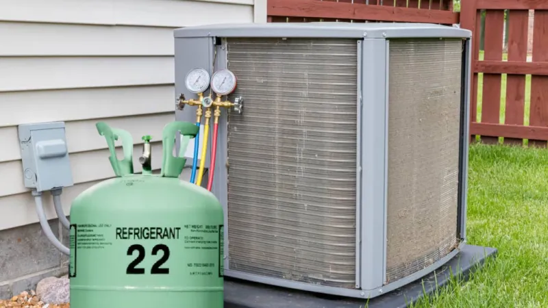 Green refrigerant 22 tank connected to an outdoor air conditioning unit near a house with red wooden fence.