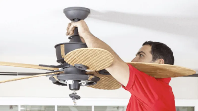 Man in red shirt installing or repairing a ceiling fan with wooden blades inside a bright room.