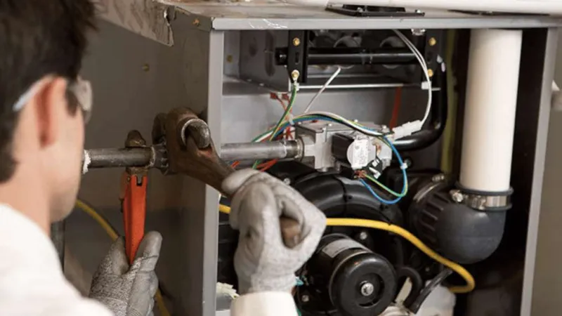Technician wearing gloves and safety glasses repairing HVAC unit using pipe wrench on indoor mechanical equipment.