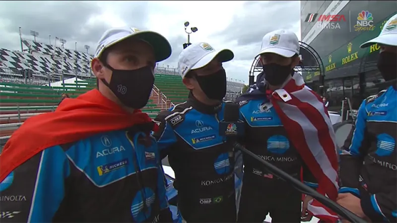 Three masked race car drivers wearing Acura suits and caps during an interview at the racetrack with flags draped over shoulders