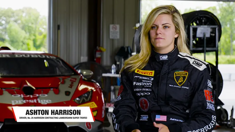 Female race car driver Ashton Harrison in black racing suit standing by a red Lamborghini race car in garage.