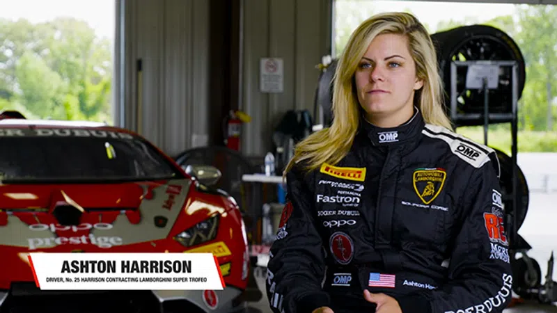 Female race car driver Ashton Harrison in black racing suit standing by a red Lamborghini race car in garage.