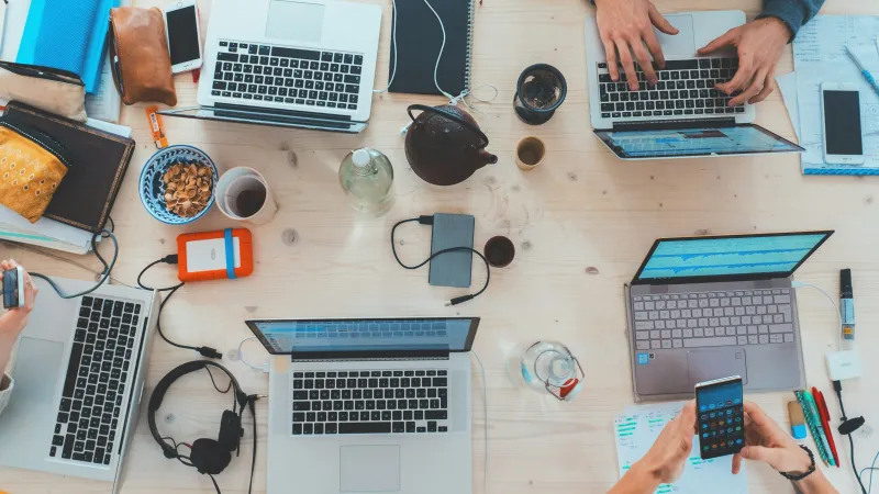 Top view of a wooden desk with laptops, notebooks, coffee, headphones, and people working collaboratively