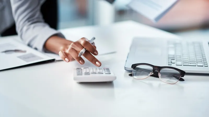 Person using calculator and laptop with glasses on a white desk in a bright office setting