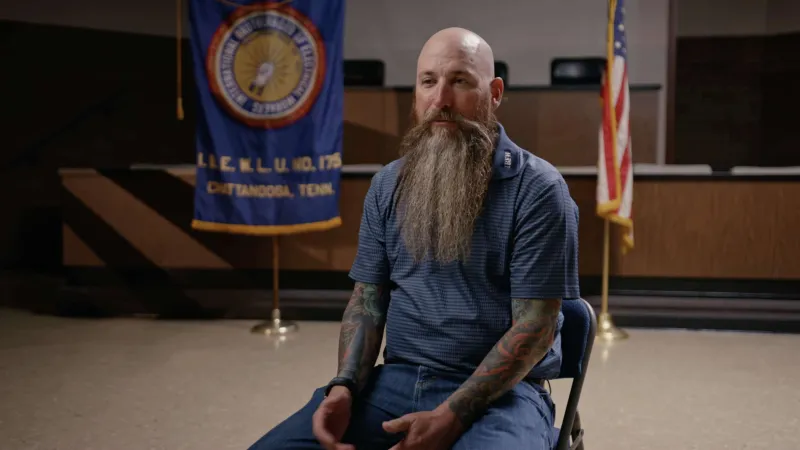 Bearded man with tattoos sitting on a chair in a meeting room with flags in the background.
