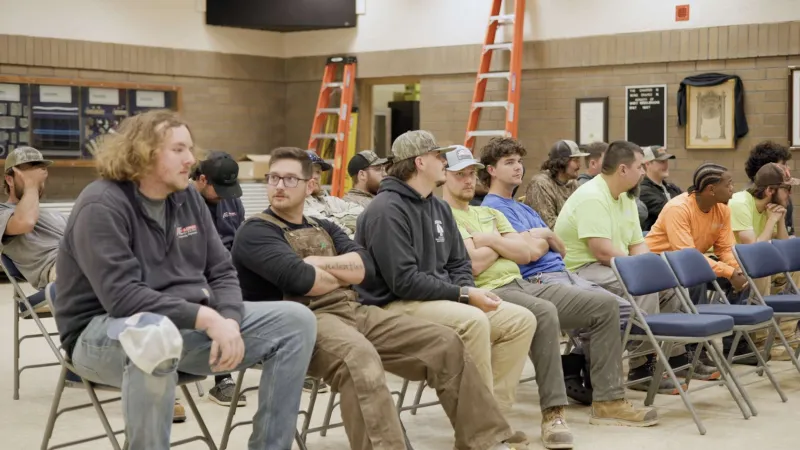 Group of men sitting on folding chairs indoors, some wearing work clothes and caps, waiting attentively.