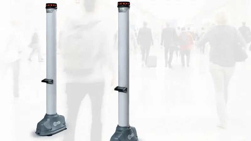 Two tall, gray security sensor pillars with red lights on top in a busy airport terminal background.