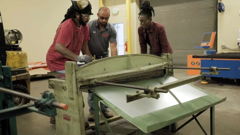 Three people operate a large metal sheet cutting machine in an industrial workshop setting.