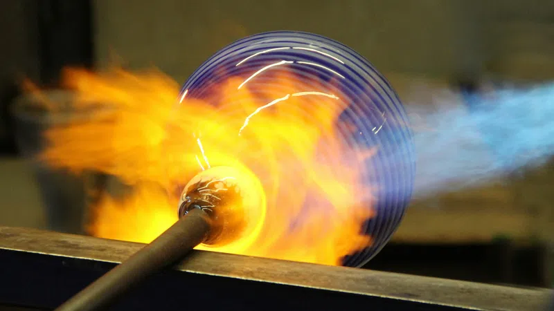 Glassblowing process with molten glass glowing orange and blue flame heating the glass on a metal rod.