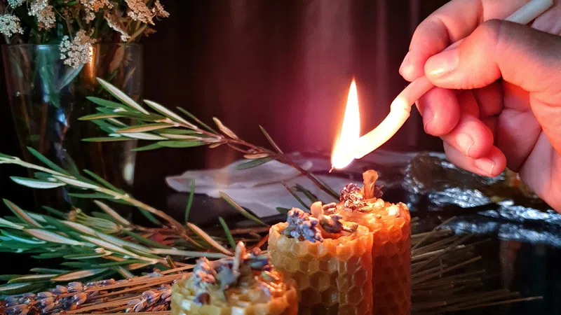 Hand lighting three textured beeswax candles with dried flowers and green foliage in the background.