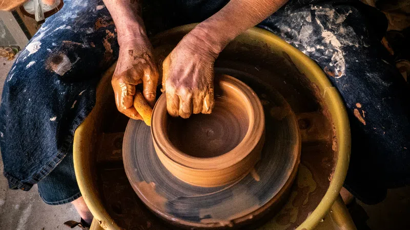 Potter shaping clay on a spinning wheel, hands covered in wet clay, crafting a round pot in a workshop.