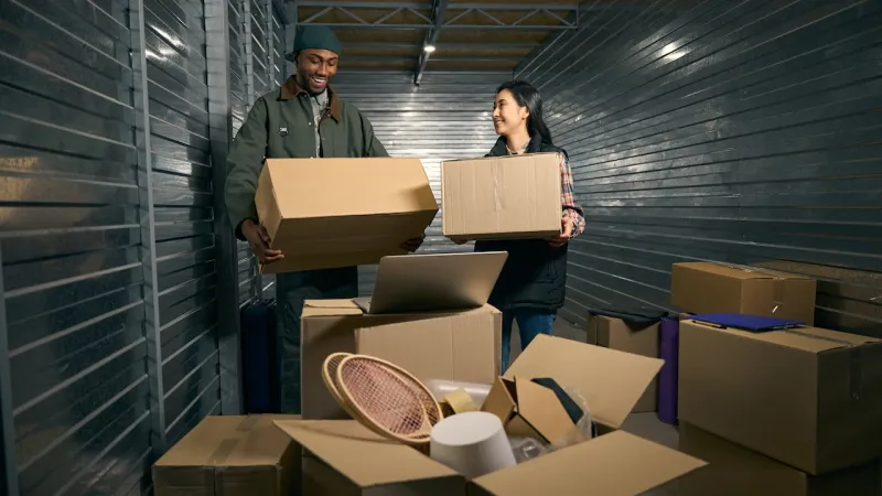 Two people smiling while unpacking boxes in a storage unit, surrounded by cardboard boxes.