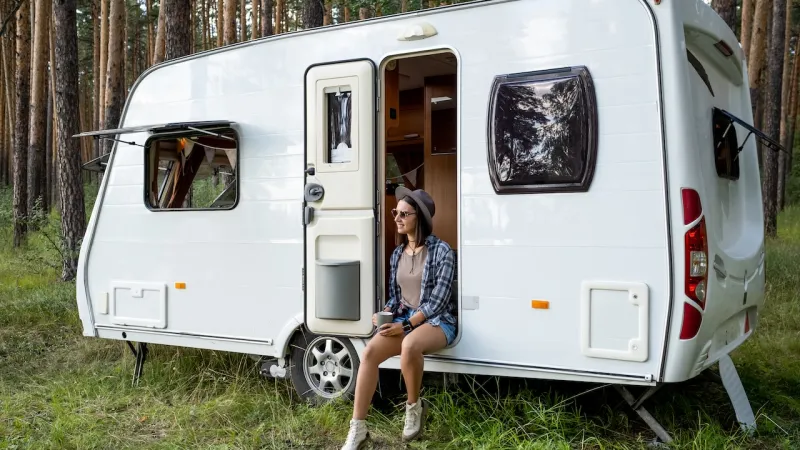 Woman enjoying nature beside a modern caravan in a forest setting.
