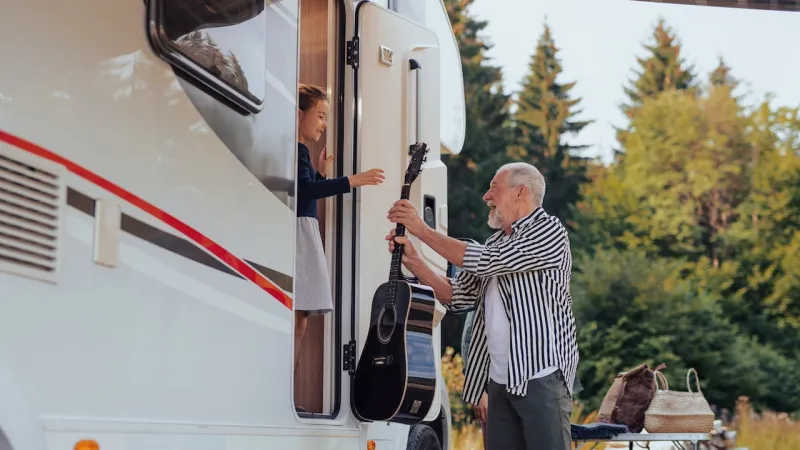 Older man hands a black acoustic guitar to a smiling girl inside a camper van with greenery in background