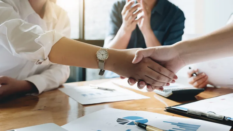 Two professionals shaking hands over business charts and laptops in a meeting room with natural light.