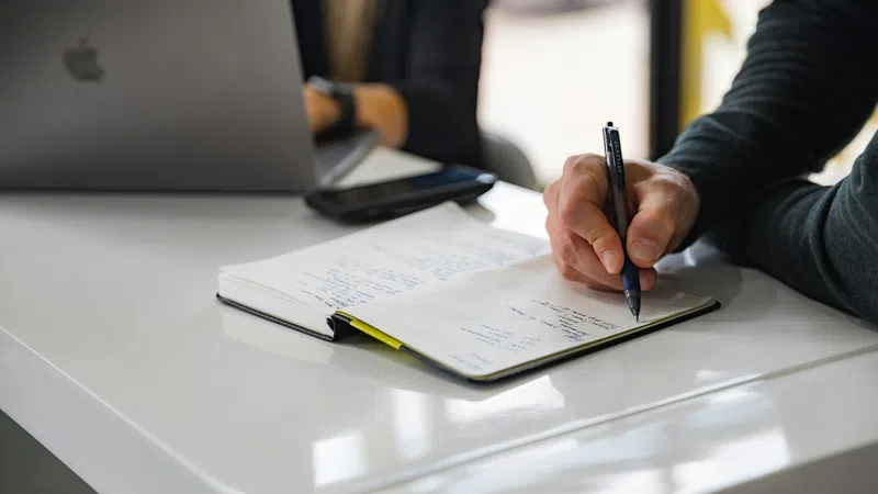 Person writing notes in a notebook at a desk with a laptop and phone nearby.