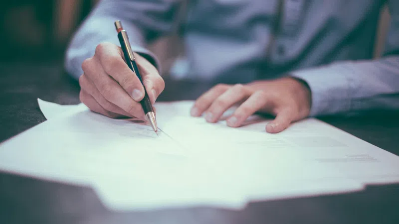 Close-up of a person writing on papers with a pen at a table, wearing a light shirt.