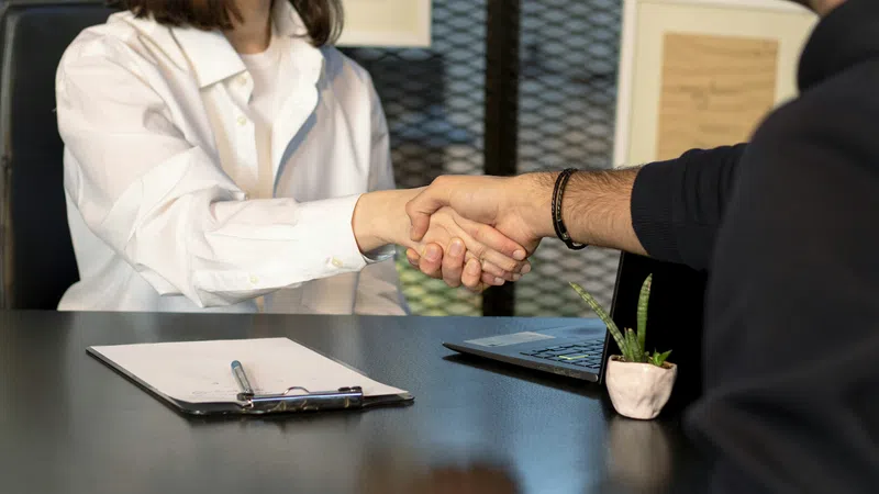 Two people shaking hands over a desk with a clipboard, pen, laptop, and small plant in an office setting.