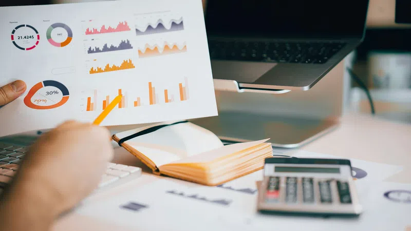 Person analyzing colorful business charts and graphs with a calculator and laptop nearby on desk.