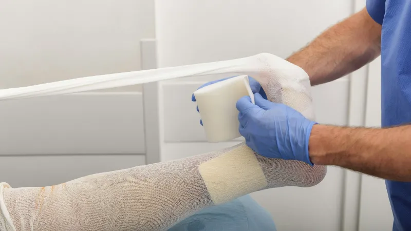 Medical professional applies a white compression bandage to a patient's leg with protective blue gloves in a clinical setting.