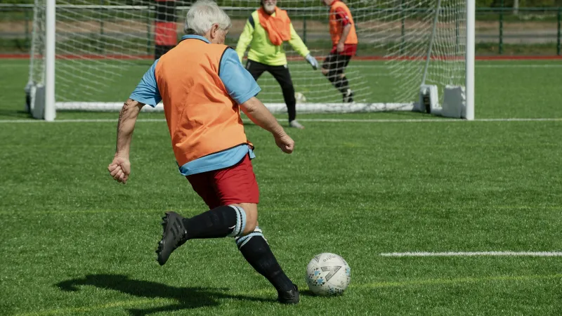 Senior man in orange vest kicking soccer ball toward goal with goalkeeper and defender in background on green field