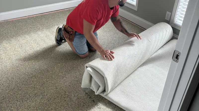 Man in red shirt kneeling and unrolling carpet padding in a sunlit empty room with gray walls and large windows.