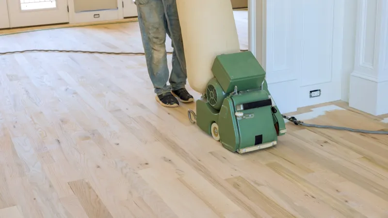Worker using a floor sanding machine to refinish a wooden floor in a bright room with white walls.