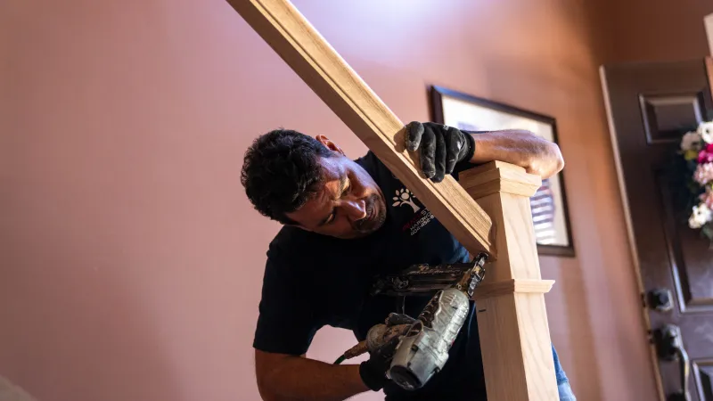 Man using nail gun to install wooden handrail on staircase inside a house with brown walls.