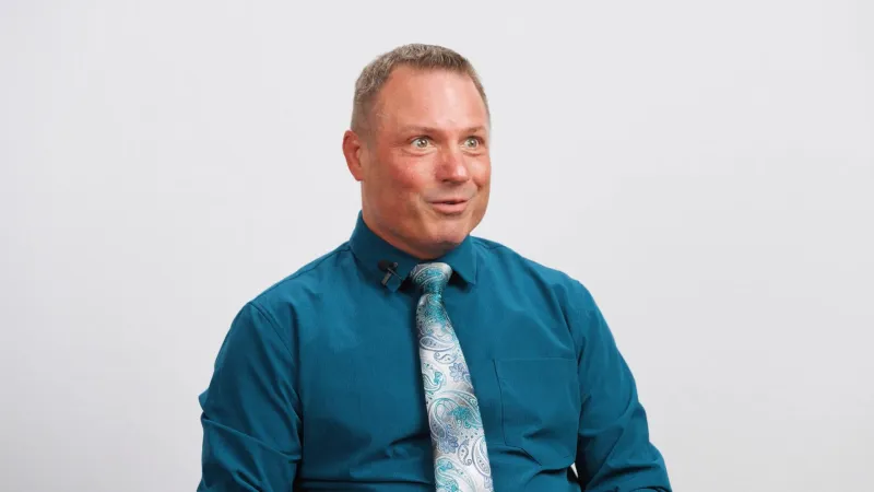 Man in teal dress shirt and paisley tie smiling against a plain white background.