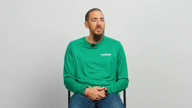 Man in green PestMaster shirt sitting on chair against a plain light gray background speaking.