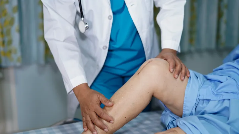 Doctor examining elderly patient's leg in a clinical setting, focusing on knee and lower limb assessment.