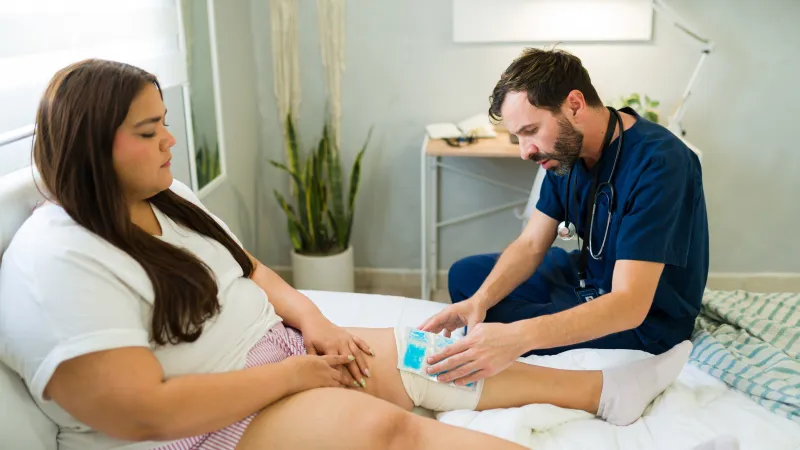 A nurse applies an ice pack to a patient's injured knee in a clinical setting with medical equipment nearby