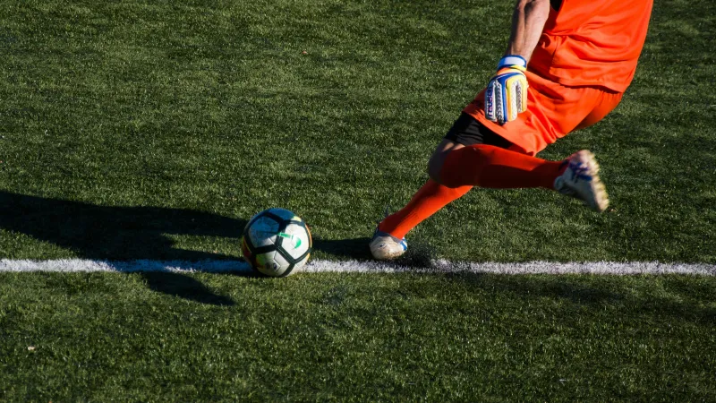 Soccer goalkeeper in orange kit kicking a ball on a green field near the sideline.