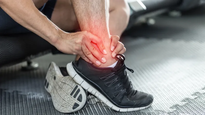 Runner holding painful ankle with red highlight, wearing black sports shoes on gym mat during workout rest.