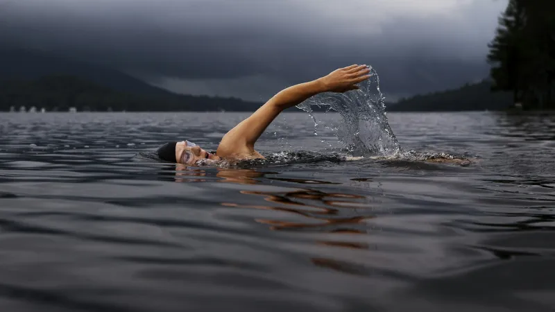 Swimmer in goggles swimming freestyle in dark, calm lake under cloudy, moody sky near forested shore.