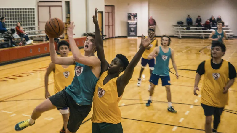 High school basketball players in blue and yellow jerseys compete for the ball during a fast-paced game in a gym.