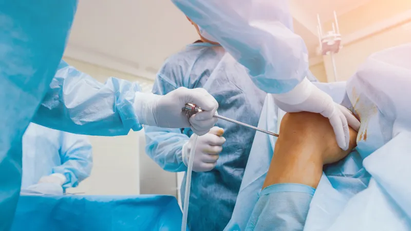 Surgeon performing arthroscopic knee surgery on patient's bent leg in sterile operating room environment