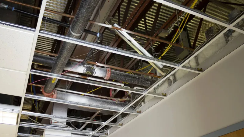 Ceiling with removed tiles exposing metal pipes, wiring, and open grid framework in an office or commercial space.