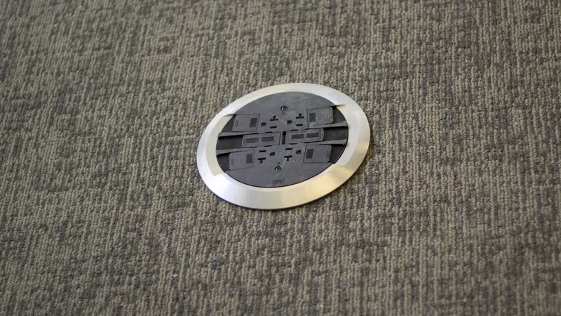 Round metal floor socket with multiple electrical outlets embedded in gray carpeted floor.