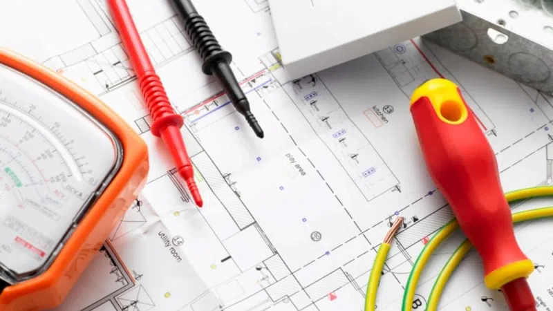 Electrical tools including a multimeter, screwdriver, and wiring on top of a detailed building circuit diagram blueprint.