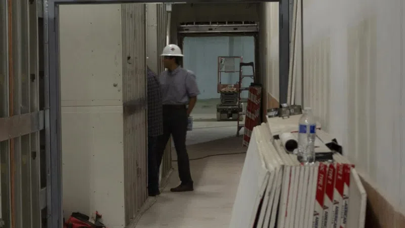 Two construction workers with hard hats inspecting drywall installation in an unfinished building corridor.