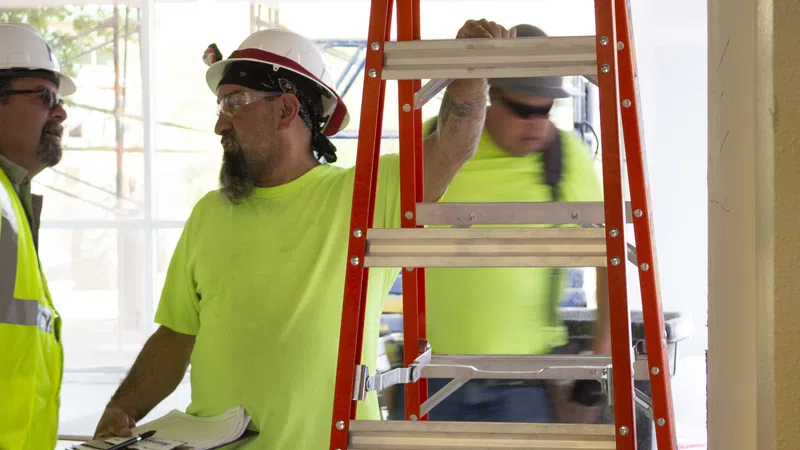 Construction workers in safety gear discussing plans at a building site with a red ladder in the foreground