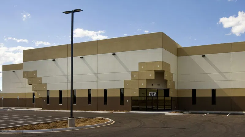 Modern beige and brown industrial warehouse with large glass entrance and empty parking lot under blue sky.